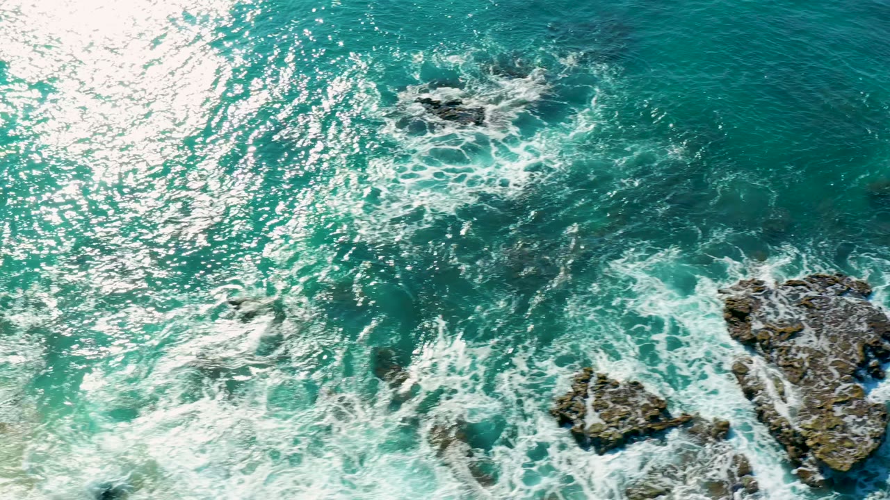 A larger dark rock sits near the right edge while white surf fans across turquoise ocean at La Fortuna, Baja California Sur, Mexico, with no visible people.