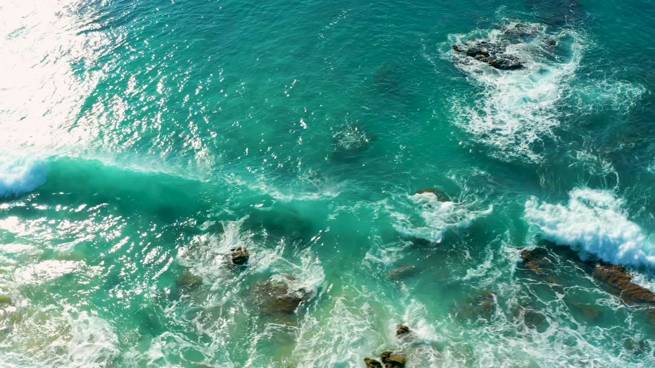 Top-down view of white foam spreading near dark rocky outcrops in turquoise water at La Fortuna, Baja California Sur, Mexico, with no visible people.