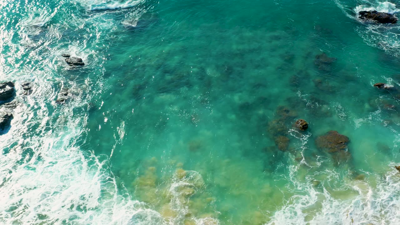 Aerial-looking frame from La Fortuna East Cape showing white foam spread through turquoise water above scattered submerged rocks, with no visible people.