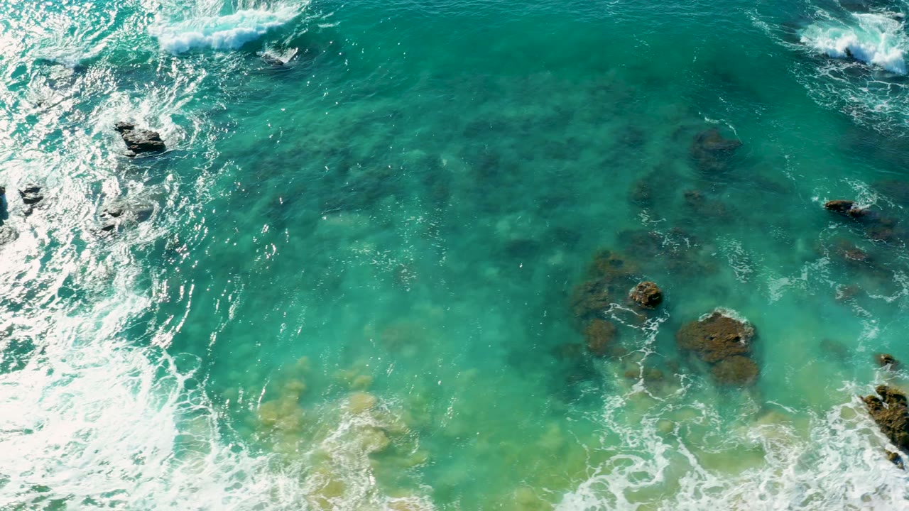 Overhead surf foam beside clear teal water and underwater rocks.