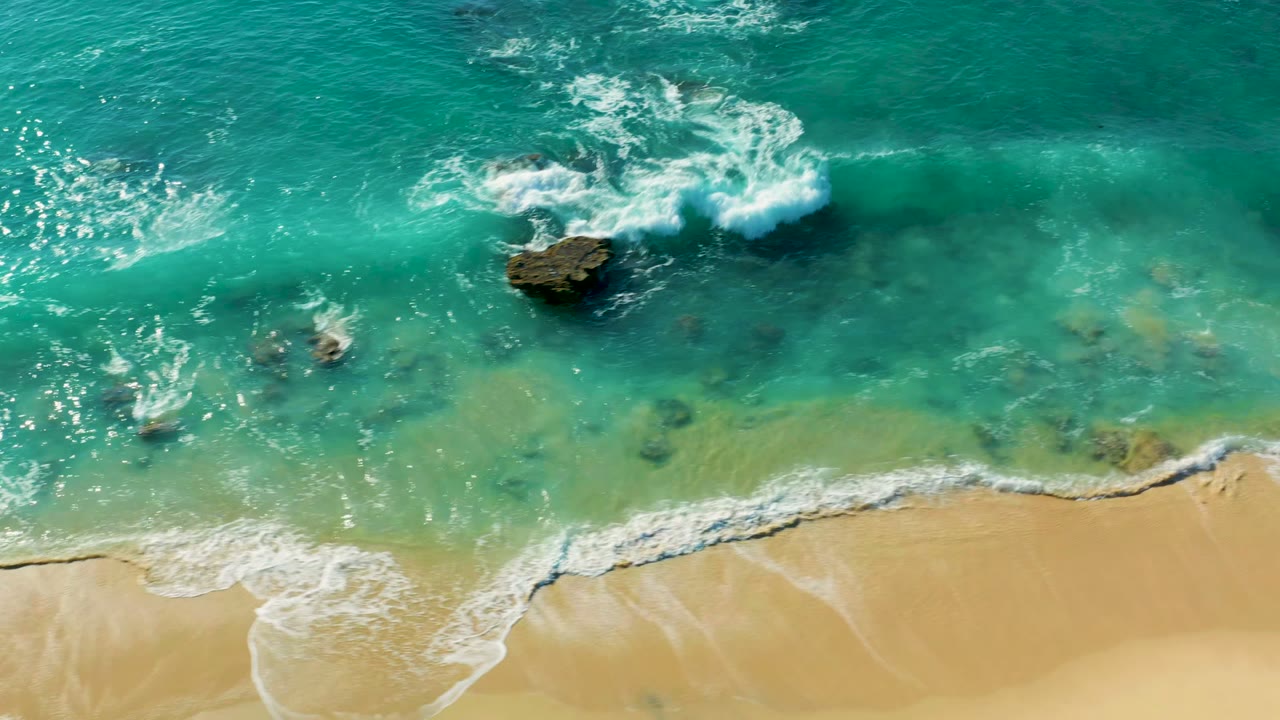 Top-down aerial view of turquoise water at La Fortuna, Baja California Sur, with white surf forming around scattered rocks and pale sand along the lower edge, no visible people.