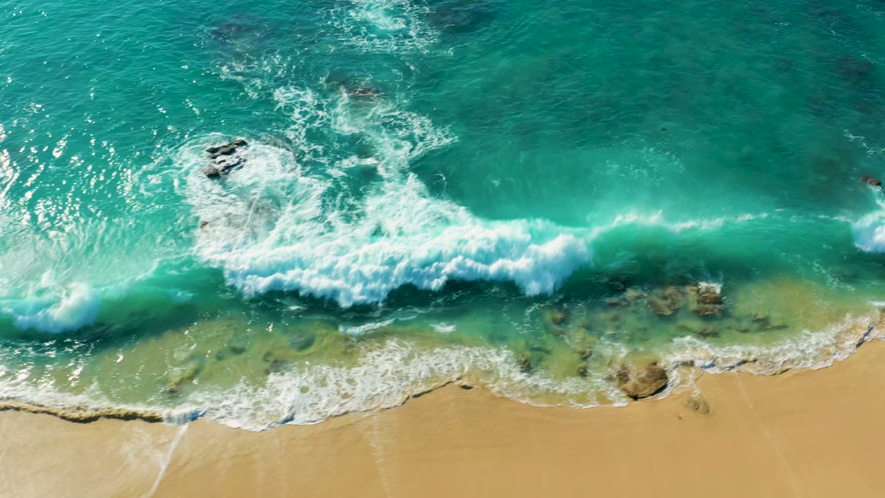Top-down view of La Fortuna surf with white foam bursting around a dark rock, green-blue shallow water below it, and smooth sand at the bottom of frame, no visible people.