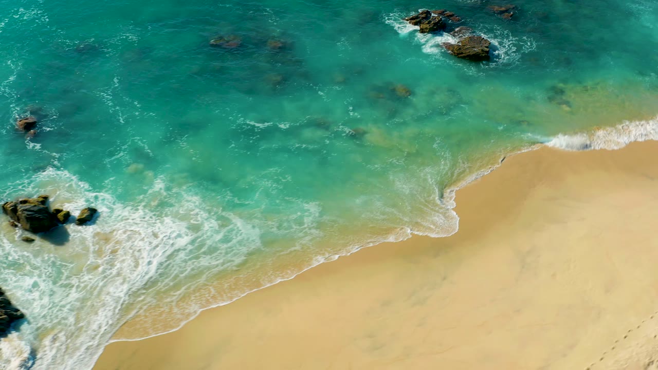 Overhead frame from La Fortuna, Baja California Sur showing turquoise surf washing onto pale sand, dark rocks in the water, and no visible people.