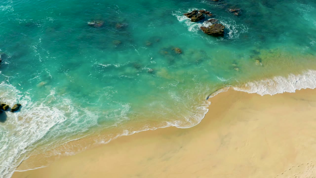 Aerial beach view with foam curling along the sand and rocks offshore.