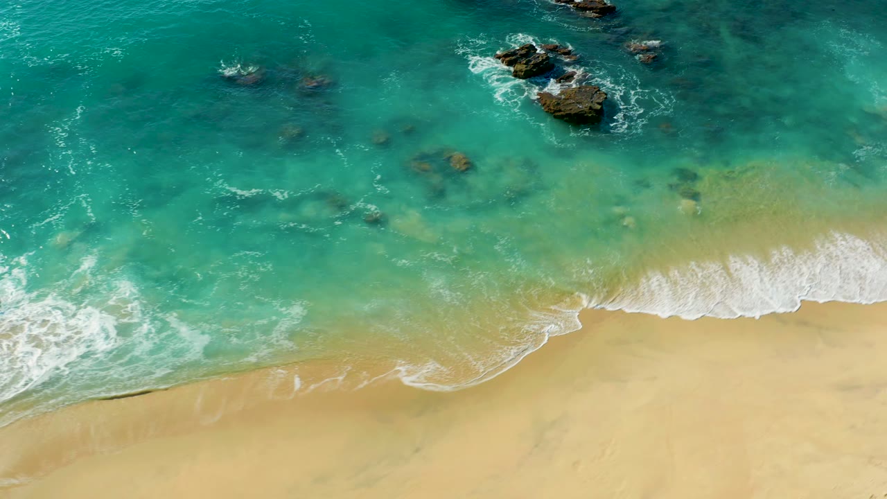 Overhead frame from La Fortuna, Baja California Sur showing a broader sandy lower edge, white foam along the right shoreline, scattered offshore rocks, and no visible people.