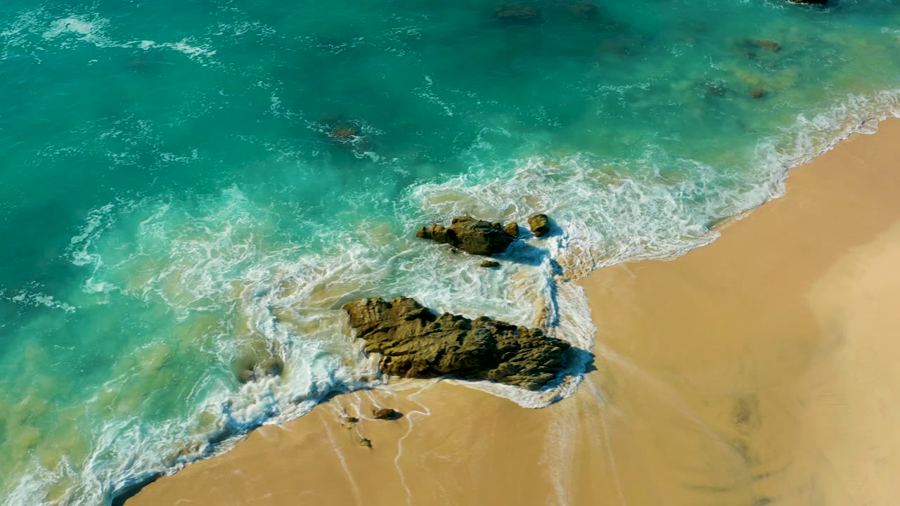 At 0:00, an overhead view in La Fortuna, Baja California Sur, shows turquoise water and white foam wrapping around a dark rock cluster on pale sand with no visible people.