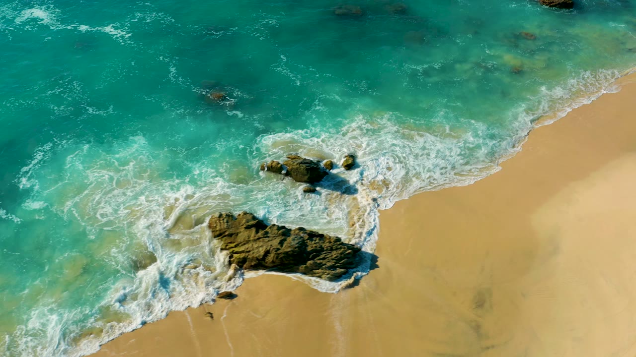Foamy wave water spreads around rocks beside a pale sandy shoreline.