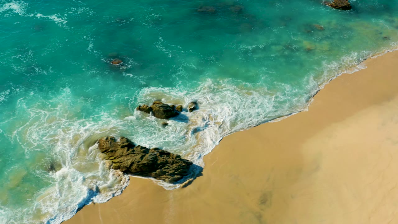 At 0:02, the aerial view shows surf curling around the larger dark rock in La Fortuna, Baja California Sur, with foam tracing the beach edge and no visible people.