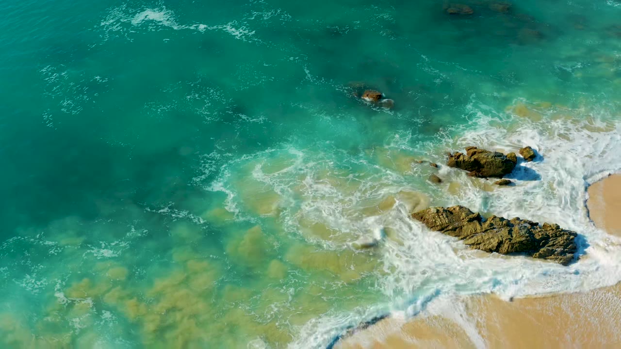 Overhead frame at La Fortuna, Baja California Sur showing turquoise water and white foam around dark rocks beside tan sand, with no visible people.