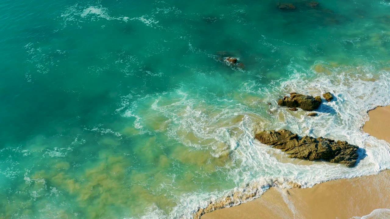 White foam spreads over shallow water near beach rocks.