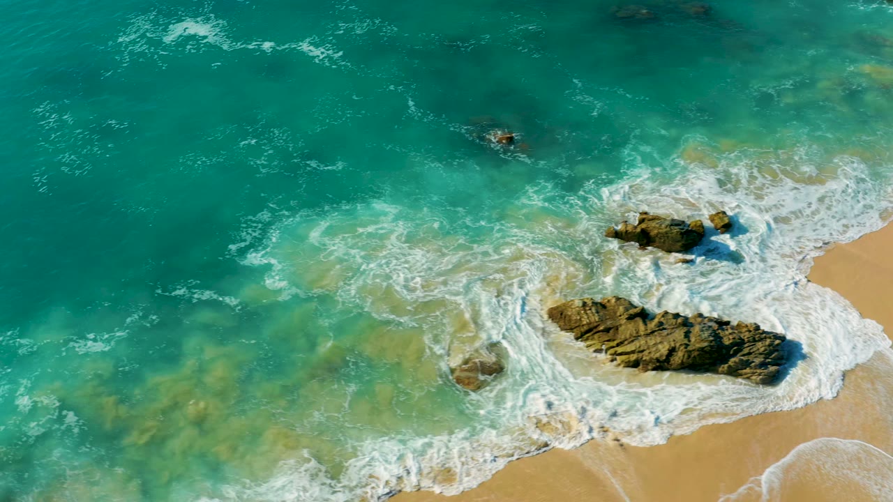 La Fortuna aerial shoreline frame with a broad turquoise water field, white wave foam curling around the central rock, and sand visible at the right, with no visible people.