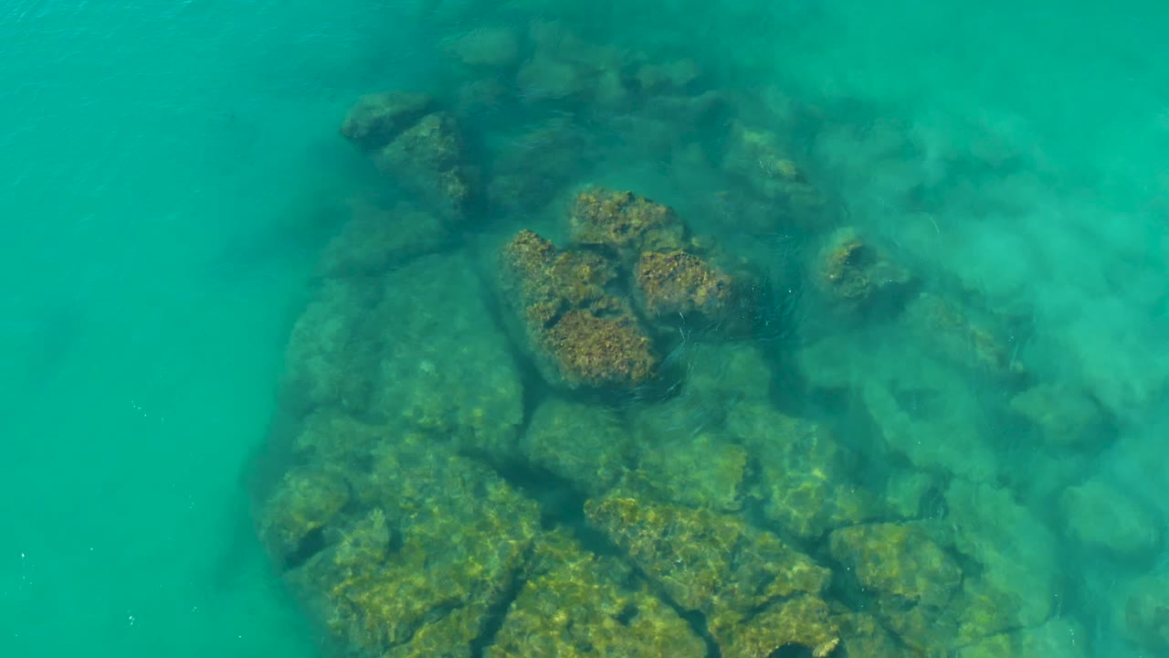At 0:02, algae-covered rocks sit below bright turquoise water in La Fortuna, with softer visibility toward the upper right and no visible people.