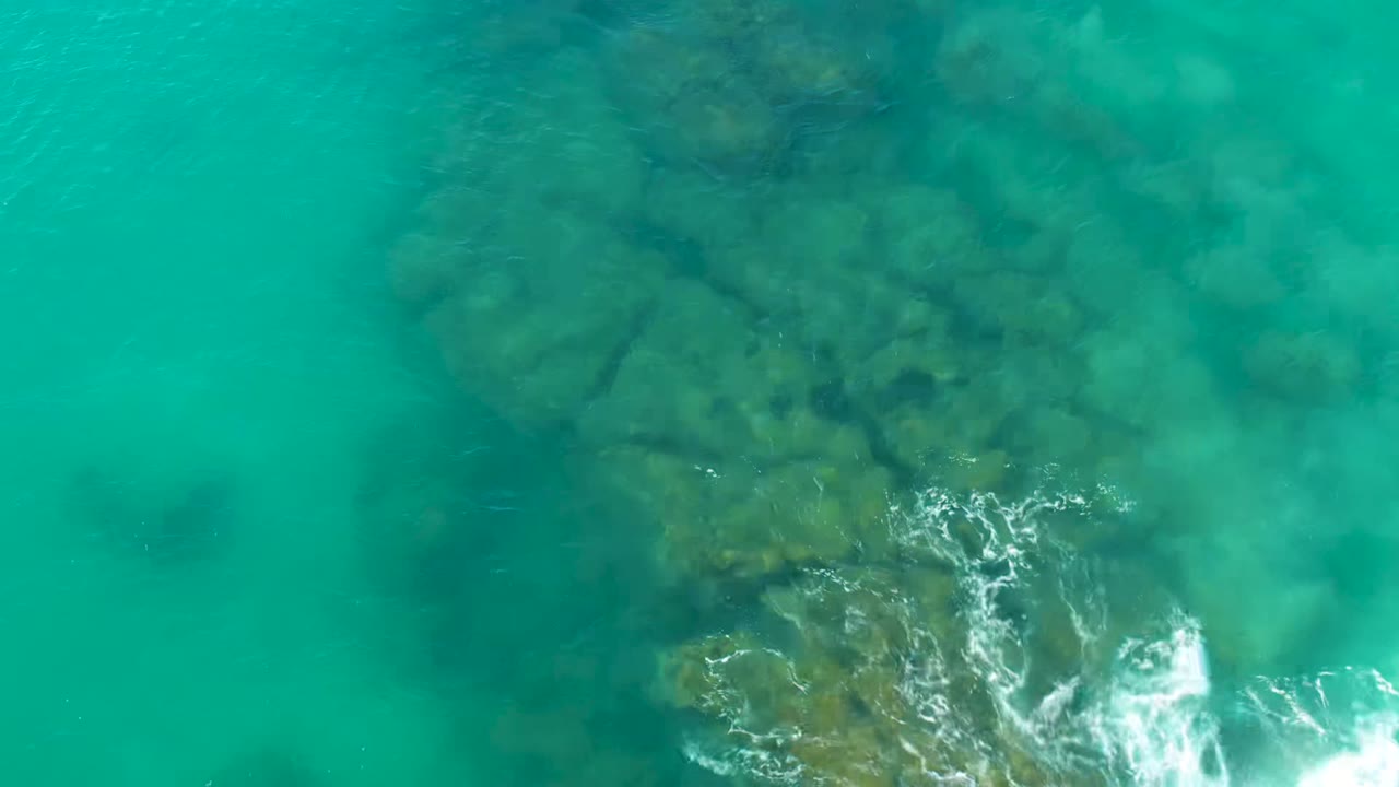 Overhead view of turquoise shallow water in La Fortuna, Baja California Sur, with dark submerged rocks and a small white foam patch near the lower edge, no visible people.