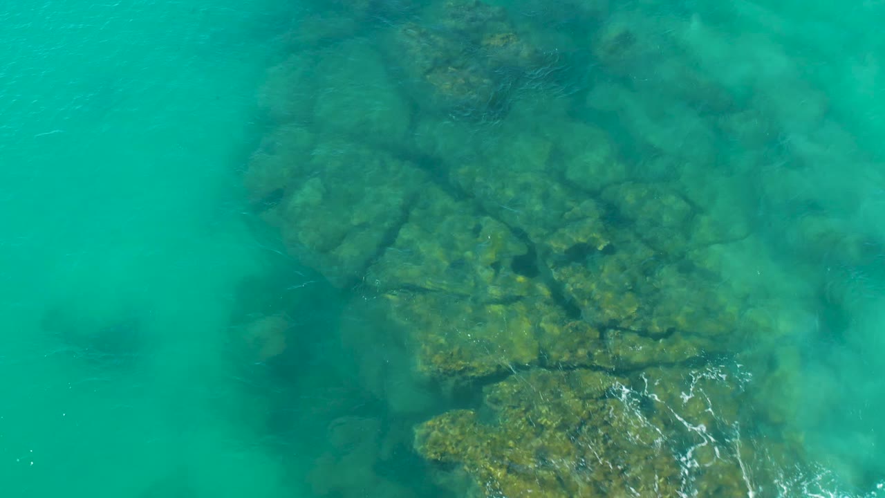 Aerial-looking view in La Fortuna, Baja California Sur, with rounded submerged rocks beneath turquoise water and a light foam streak at the lower right, no visible people.