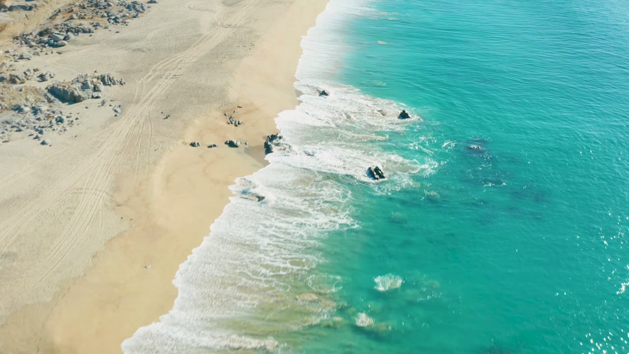 Aerial view over La Fortuna, Baja California Sur, with turquoise water rolling onto pale sand, dark rocks in the surf, faint beach tracks, and no visible people.