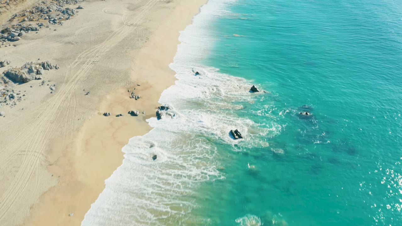 Aerial frame from La Fortuna showing a diagonal foam line where turquoise ocean meets pale beach, with several dark rocks near the breaking waves and no visible people.