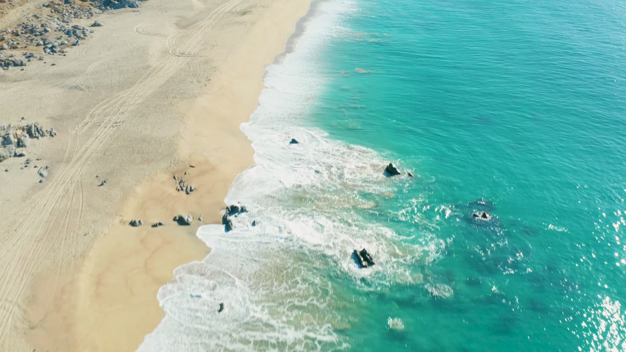 Aerial view of La Fortuna shoreline with bright turquoise water filling the right side, pale sand on the left, white surf wrapping around offshore rocks, and no visible people.
