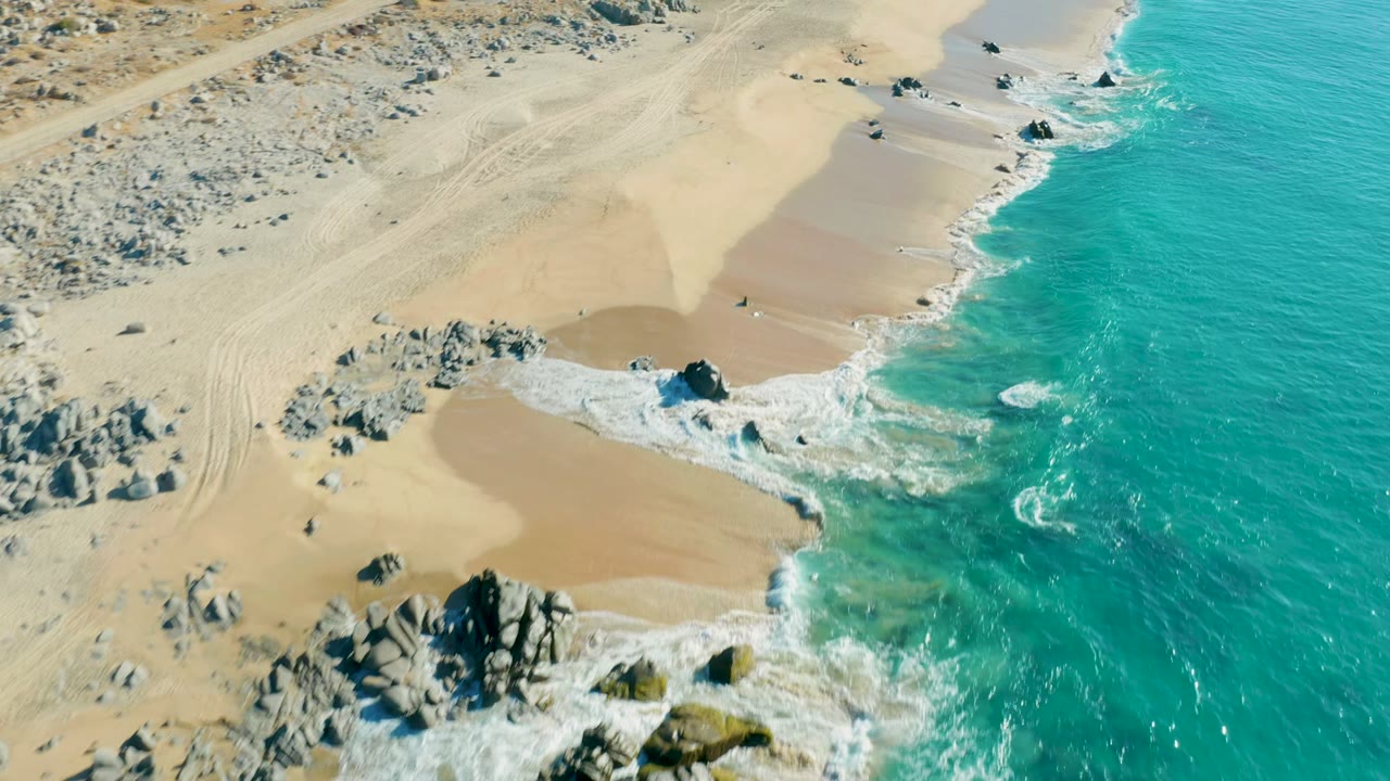 Aerial frame over La Fortuna, Baja California Sur, showing turquoise ocean on the right, white foam around dark rocks, tan sand to the left, and no visible people.