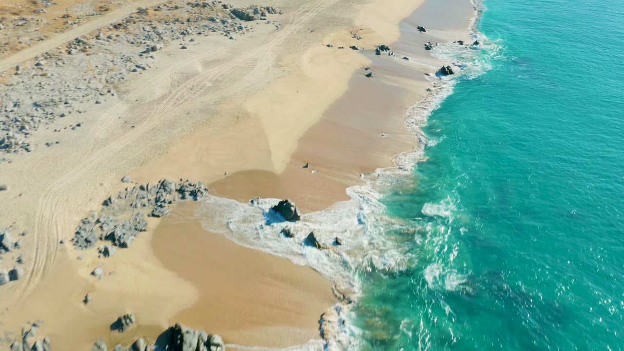 High aerial shoreline view with waves breaking beside dark rocks on tan sand.