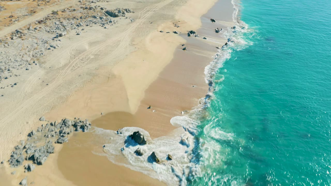 Aerial frame over La Fortuna beach with the ocean occupying the right half, white surf curling into a sandy cove, exposed rocks near the waterline, and no visible people.
