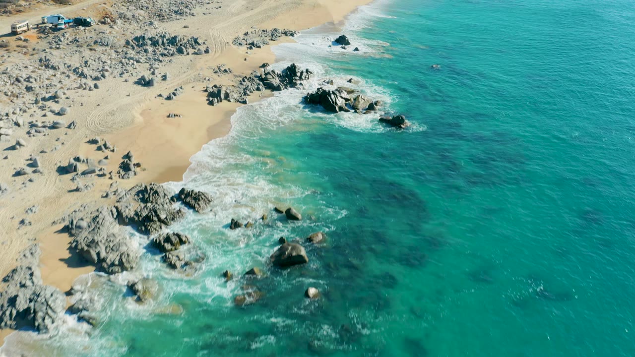 Aerial view over La Fortuna, Baja California Sur, with turquoise water meeting pale sand and gray rock outcrops while white surf wraps around the rocks, no visible people.