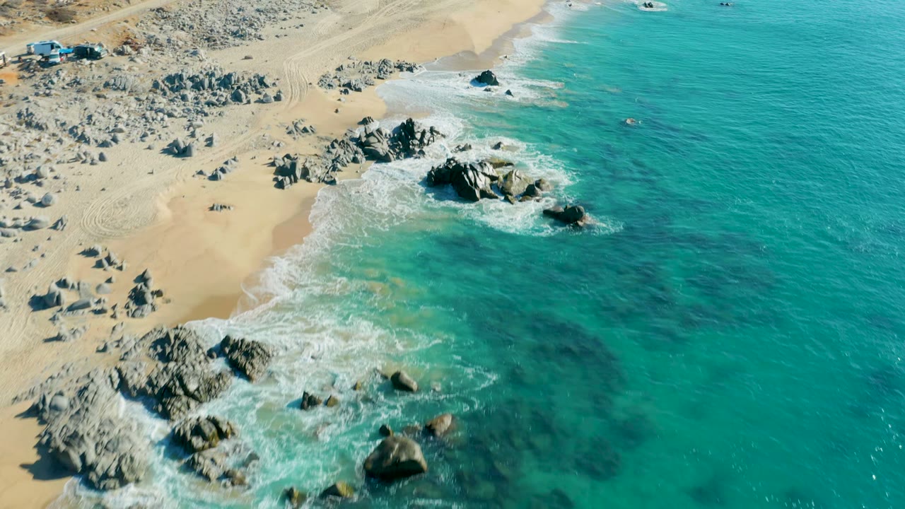 Aerial coastline view with surf foaming around rocks beside pale sand.