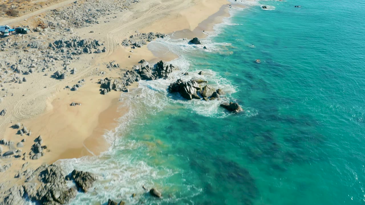 High oblique view of La Fortuna shoreline with a broad turquoise water field, white wash at the waterline, pale sand, and clusters of gray rocks inland and at the surf edge, no visible people.