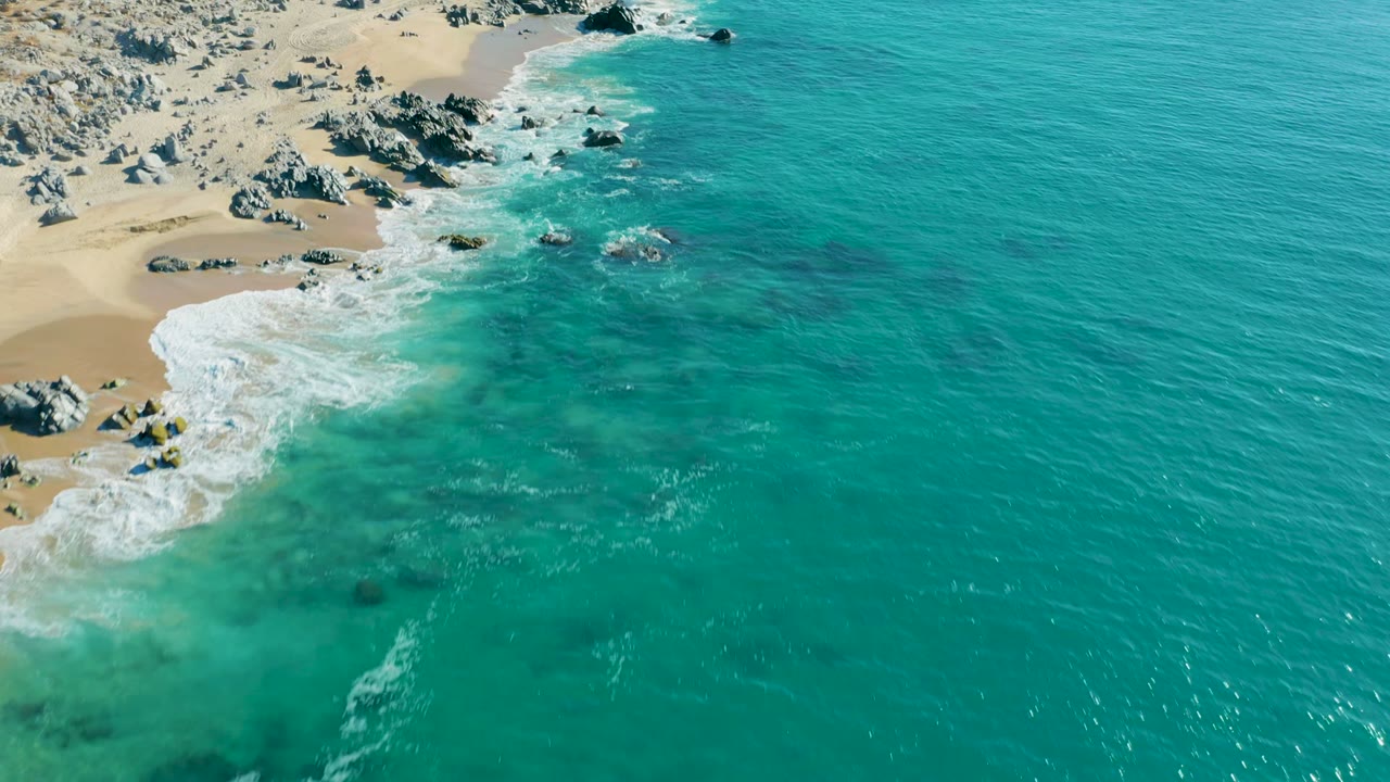 Aerial view at La Fortuna, Baja California Sur with turquoise water rolling toward a tan rocky beach, white foam along the shore, and no visible people.