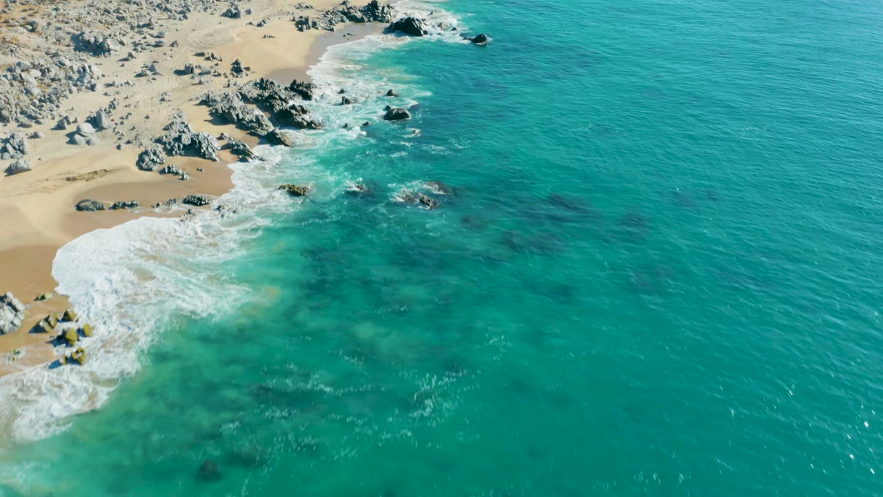 Drone view of white surf pushing into rocks on a sandy shoreline.