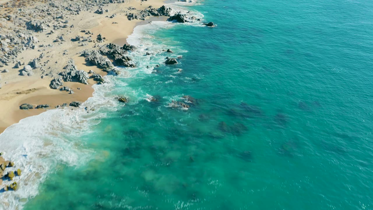 Aerial frame from La Fortuna, Baja California Sur showing clear turquoise ocean, dark submerged rock shapes, a sandy beach at left, and no visible people.
