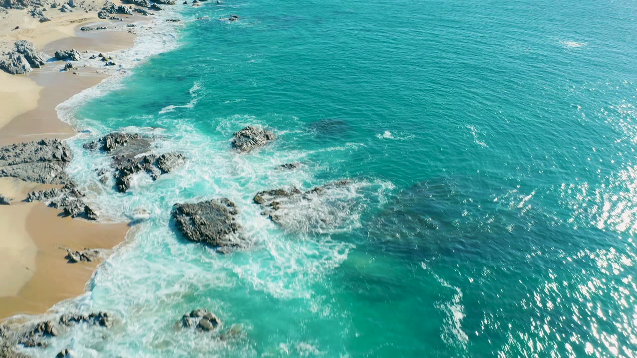 Aerial frame from La Fortuna, Baja California Sur, showing turquoise surf foaming around dark shoreline rocks with pale sand at the left edge and no visible people.