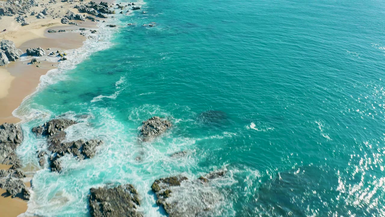 Aerial frame from La Fortuna, Baja California Sur, showing a diagonal sandy beach, clustered rocks in foamy surf, broad turquoise water to the right, and no visible people.