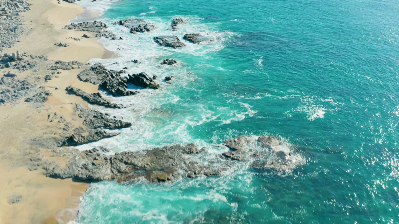 Aerial view over La Fortuna, Baja California Sur, where turquoise surf breaks around dark shoreline rocks beside pale sand, with no visible people.