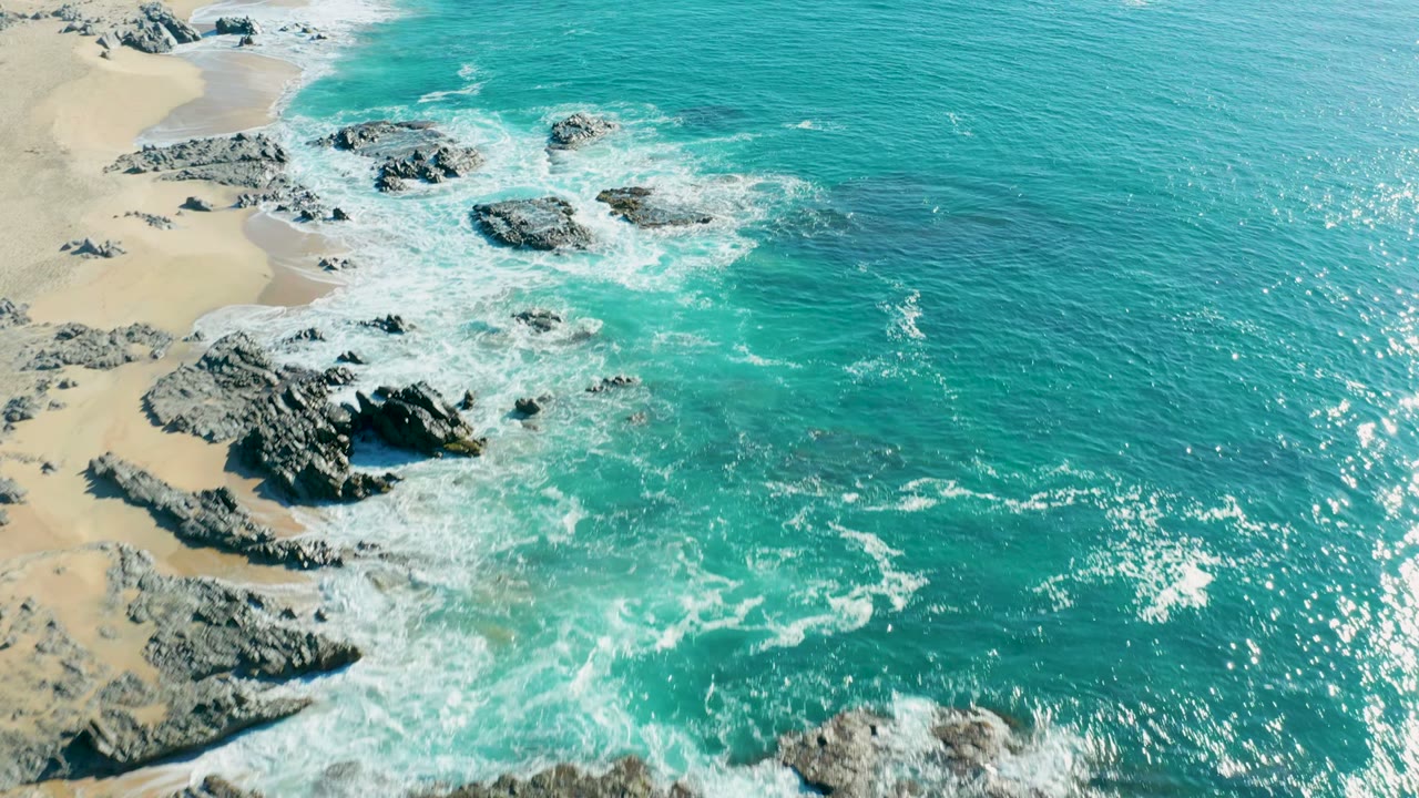 At 0:02, the aerial composition places pale sand and black rocks to the left with bright turquoise sea and sun sparkle across the right, with no visible people.