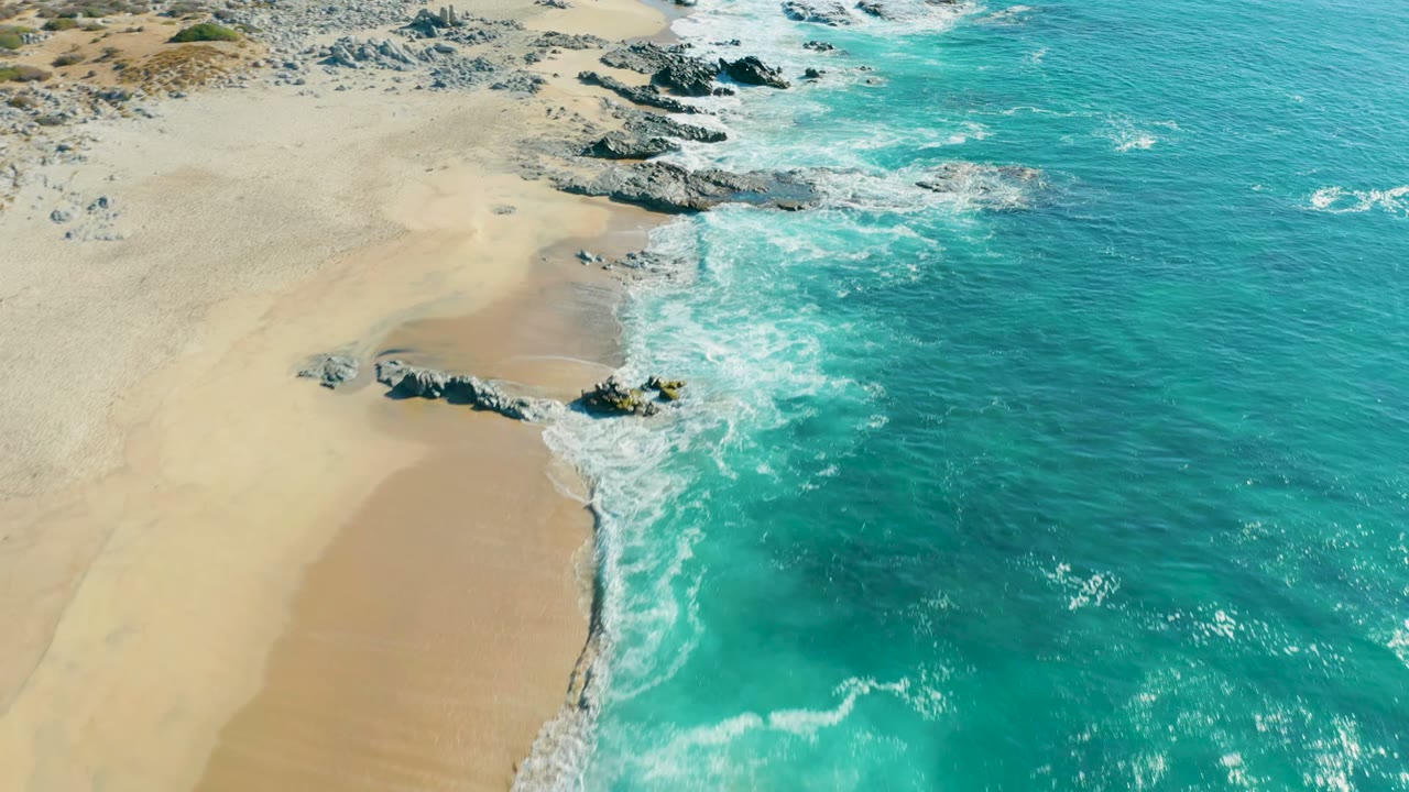 Aerial frame over La Fortuna, Baja California Sur showing turquoise water foaming against pale sand and a low rocky point, with no visible people.