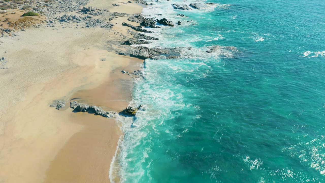 Aerial frame over La Fortuna, Baja California Sur with more white foam spreading across the waterline beside a protruding rock formation, with no visible people.