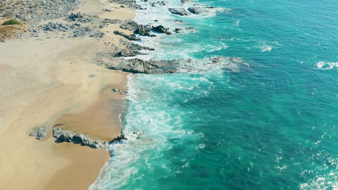 Aerial frame over La Fortuna, Baja California Sur showing the shoreline shifted farther along the rocky coast, with white surf curling against sand and no visible people.