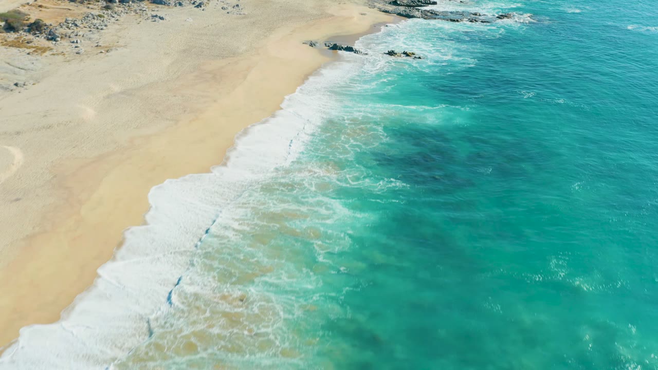 Aerial view at La Fortuna, Baja California Sur, with pale sand on the left, turquoise surf rolling in from the right, rocky coast near the top, and no visible people.