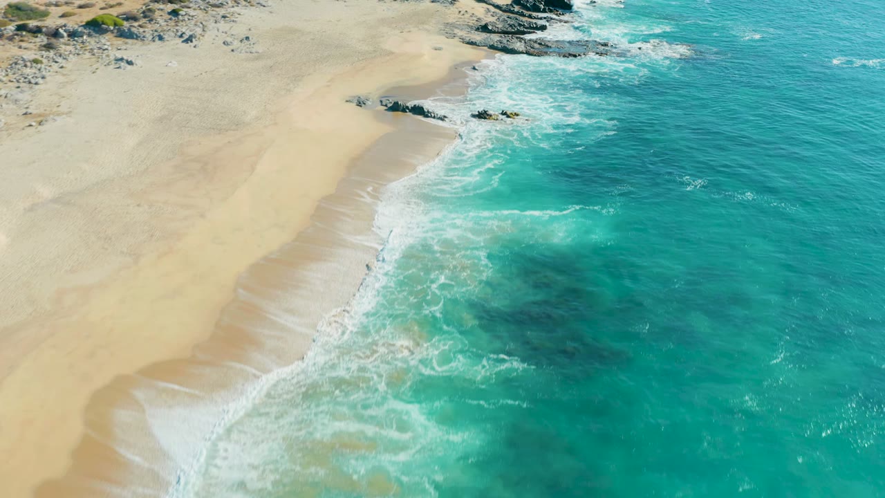 Aerial view of La Fortuna beach with more open water filling the right side, bright foam crossing the center, exposed rocks at the upper edge, and no visible people.