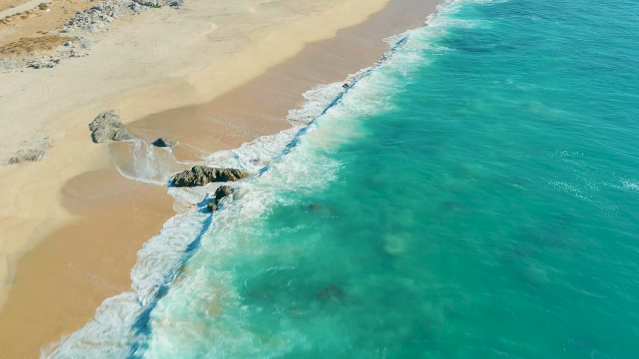 Aerial view of La Fortuna, Baja California Sur shoreline as turquoise water reaches pale sand and dark rocks at the waterline, with no visible people.