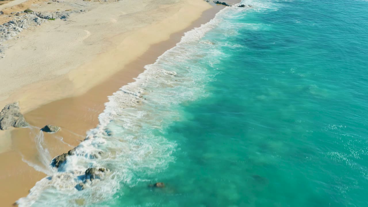 Aerial view over La Fortuna, Baja California Sur as a broken white foam line runs along the beach and turquoise shallows fill the right side, with no visible people.