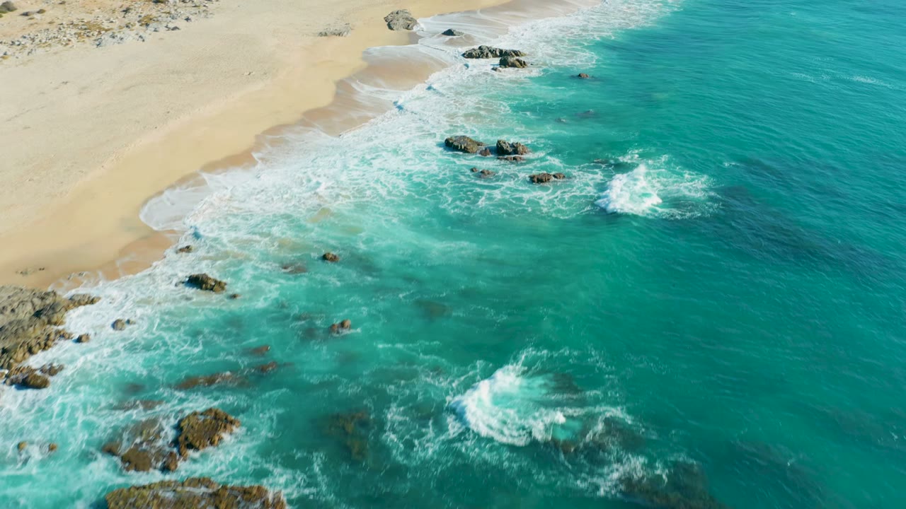 Aerial frame over La Fortuna, Baja California Sur, Mexico, showing turquoise surf around scattered rocks with pale sand at the upper edge and no visible people.