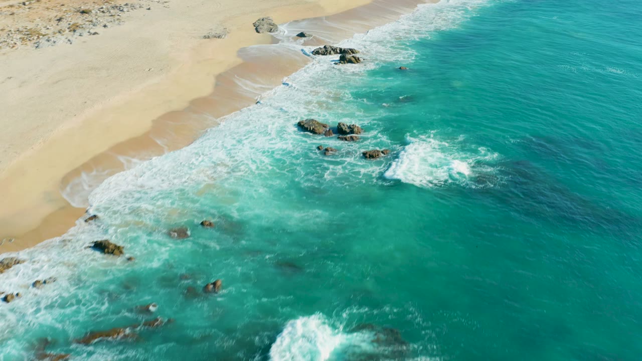 White surf spreads around dark rocks along a pale sandy shoreline