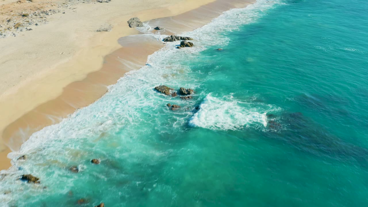 Aerial frame from La Fortuna showing a brighter patch of breaking surf near the right side, clustered rocks below the sand, turquoise water, and no visible people.