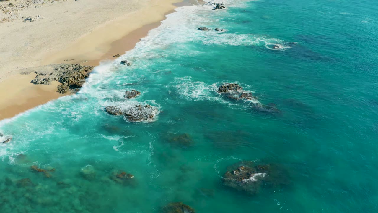 Aerial view over La Fortuna, Baja California Sur, Mexico, with turquoise surf breaking around scattered rocks beside a pale sandy beach and no visible people.