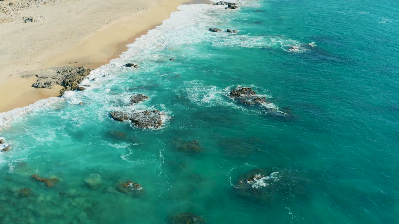 Turquoise ocean water churns around rocks near a sandy shoreline.