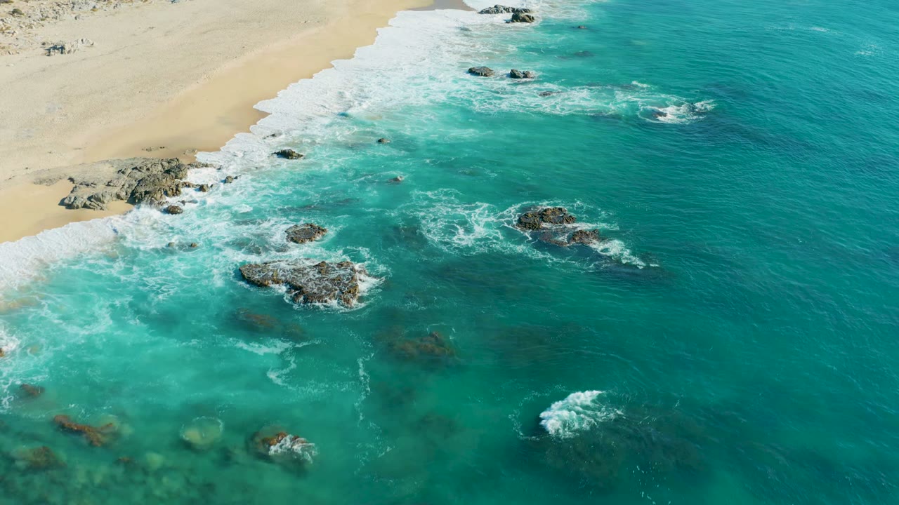Aerial view at La Fortuna with a sandy beach along the upper left, dark rock clusters in clear turquoise water, fresh white foam around the rocks, and no visible people.