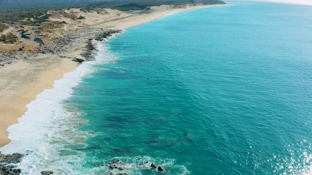 Aerial frame over La Fortuna, Baja California Sur, showing turquoise surf sweeping onto pale sand with rocks at the lower left and no visible people.