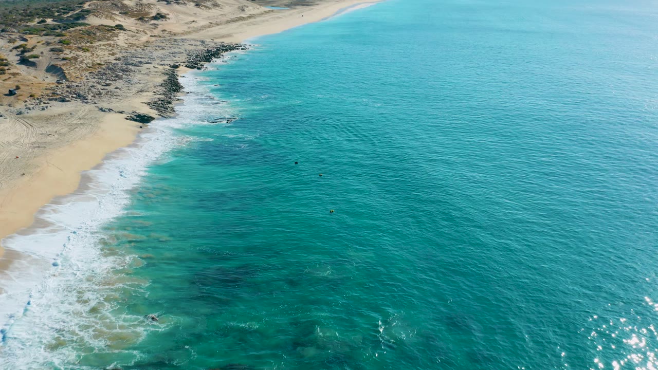 Wide aerial frame from La Fortuna with open blue-green water on the right, a breaking foam band along the beach, rocky points near the shore, and no visible people.