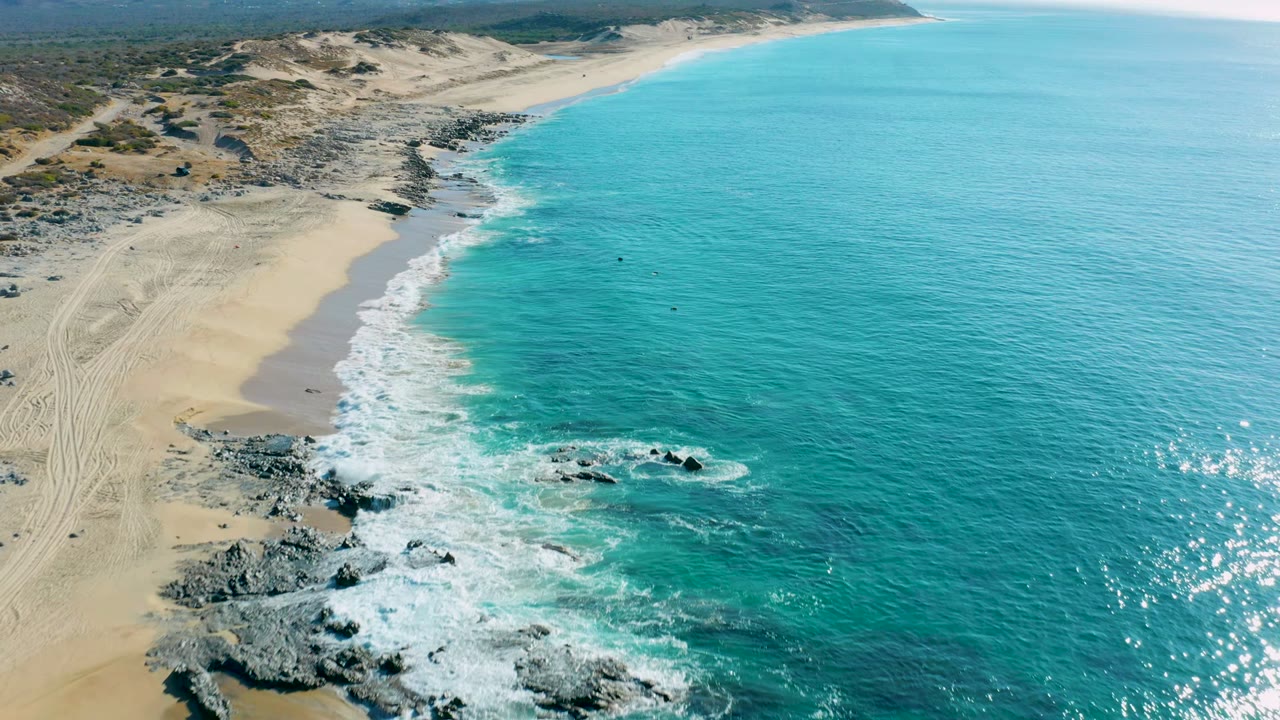 La Fortuna shoreline seen from above, with surf thickening around the central rocks, clear turquoise water to the right, and empty sand and dunes to the left, with no visible people.